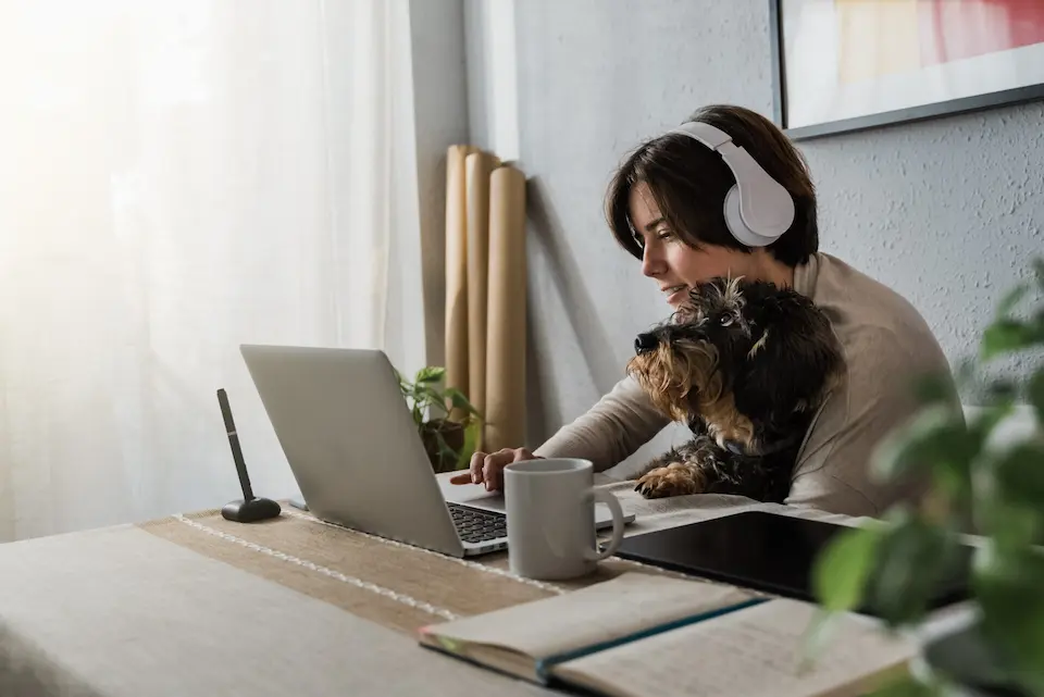 Eine Person mit Kopfhörern arbeitet an einem Tisch mit einem Laptop und einem Hund auf dem Schoß, möglicherweise in einer Online-Sitzung.