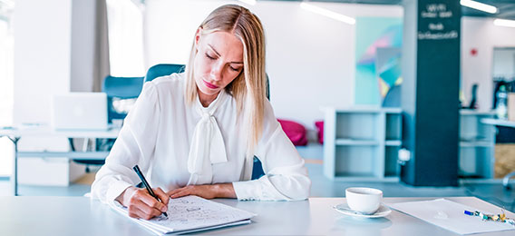 Eine Frau in einer weißen Bluse sitzt an einem Schreibtisch und schreibt in ein Notizbuch. Eine Tasse Kaffee und einige Papiere liegen auf dem Schreibtisch in einer modernen Büroumgebung.