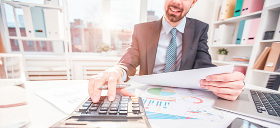 Ein Mann im Anzug verwendet einen Taschenrechner und überprüft Finanzdokumente an einem Schreibtisch mit Diagrammen, Papieren und einem Laptop in einer Büroumgebung.