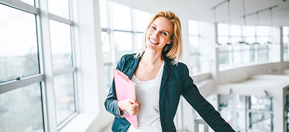 Eine Frau mit blondem Haar, einem schwarzen Blazer und einem weißen Hemd steht in einem Gebäude in der Nähe großer Fenster, hält einen rosa Ordner in der Hand und lächelt in die Kamera.