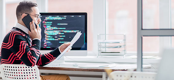 Ein Mann sitzt an einem Schreibtisch und betrachtet Dokumente, während er telefoniert. Auf dem Computermonitor hinter ihm werden bunte Codezeilen angezeigt.