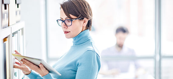 Eine Frau mit Brille und hellblauem Rollkragenpullover wählt in einem Büro eine Akte aus einem Regal. Im Hintergrund ist eine verschwommene Person zu sehen, die an einem Schreibtisch arbeitet.