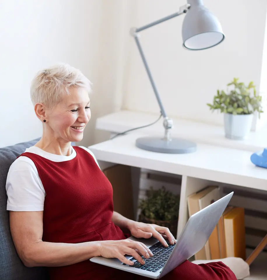Ältere Frau mit kurzen Haaren, lächelt und tippt auf einem Laptop auf einem Sofa neben einem Schreibtisch mit Lampe und Pflanzen.
