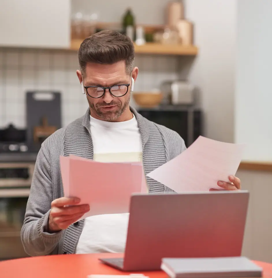 Ein Mann mit Brille und Ohrstöpseln prüft Dokumente, während er in seiner Küche an einem Laptop arbeitet.