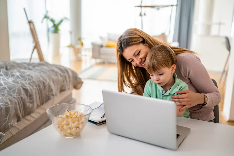 Eine Frau und ein Kind schauen gemeinsam in einem gemütlichen Wohnzimmer auf den Bildschirm eines Laptops. Auf dem Tisch steht eine Schale Popcorn.