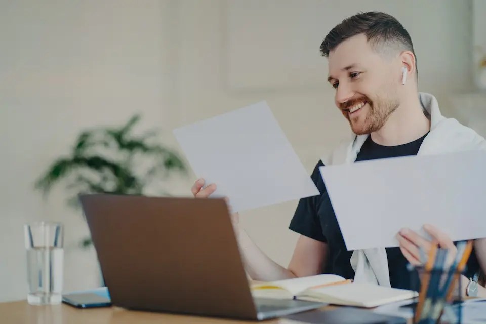Ein lächelnder Mann mit zwei Blättern Papier in der Hand, der mit einem Laptop, einem Notizbuch und einem Glas Wasser an einem Schreibtisch sitzt und seine Lernsitzung genießt.